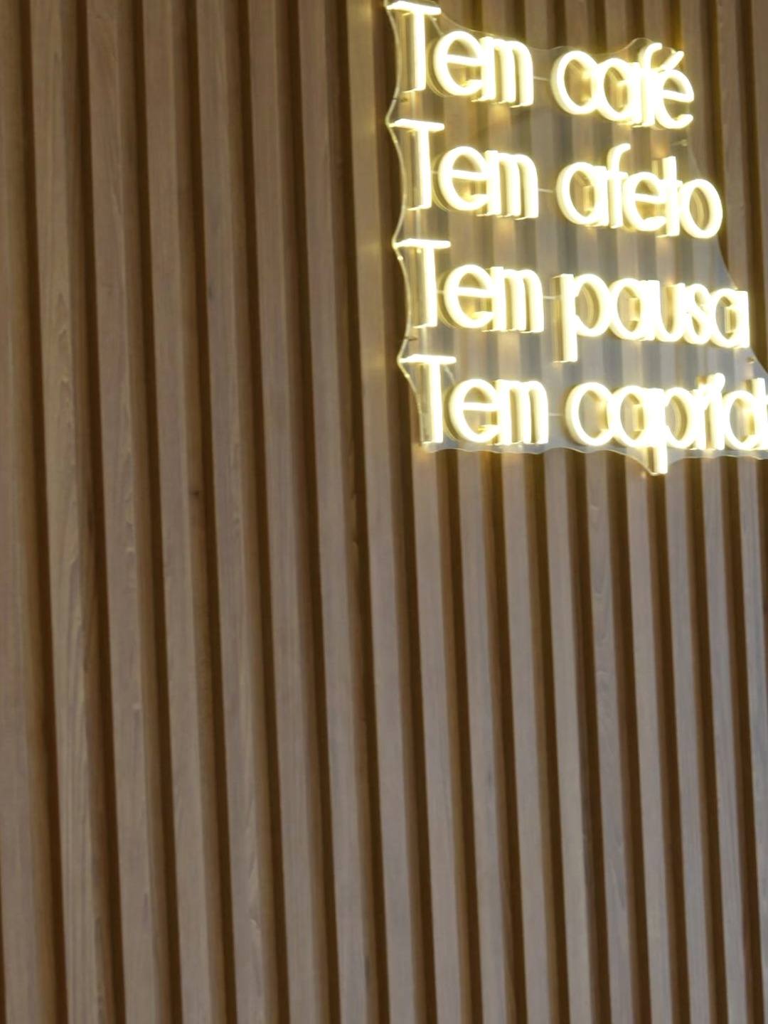 Painel ripado amadeirado com o neon "Tem café, tem afeto, tem pausa, tem capricho"